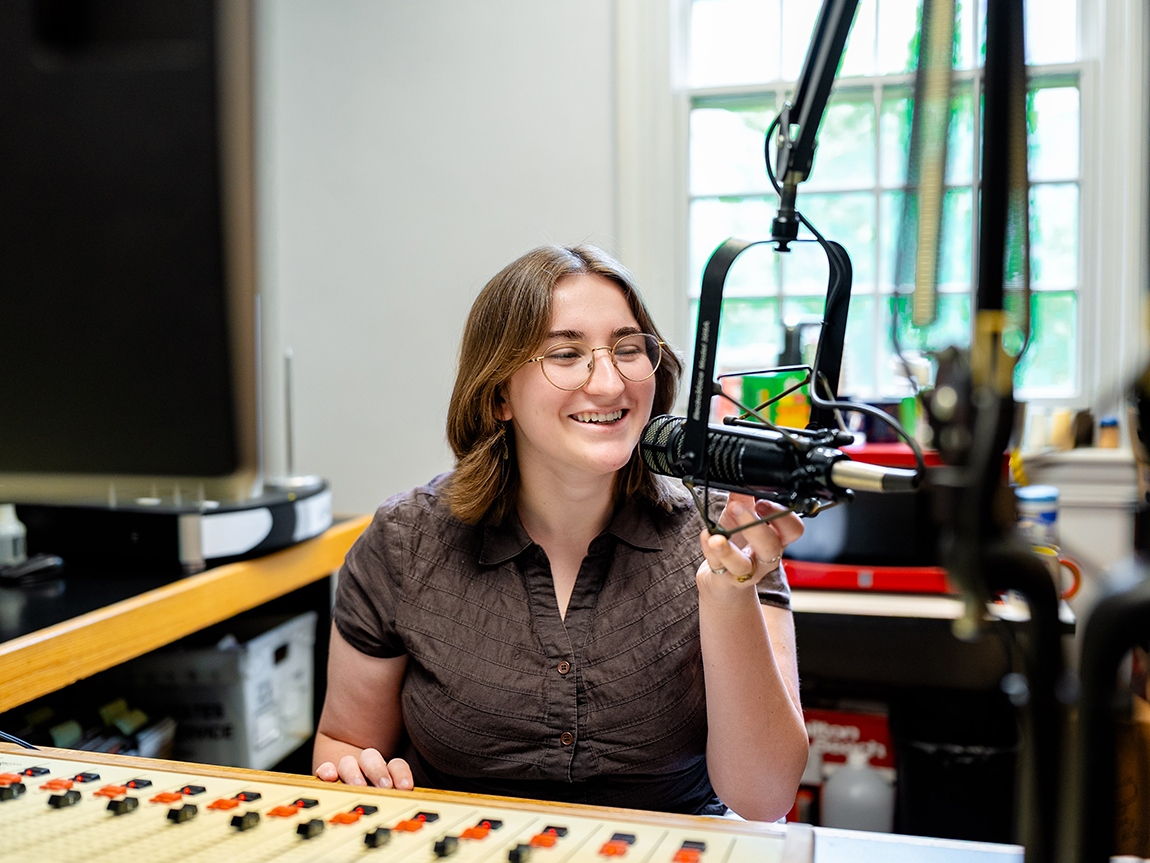 A college student in a brown shirt and glasses speaks into a microphone in a recording studio